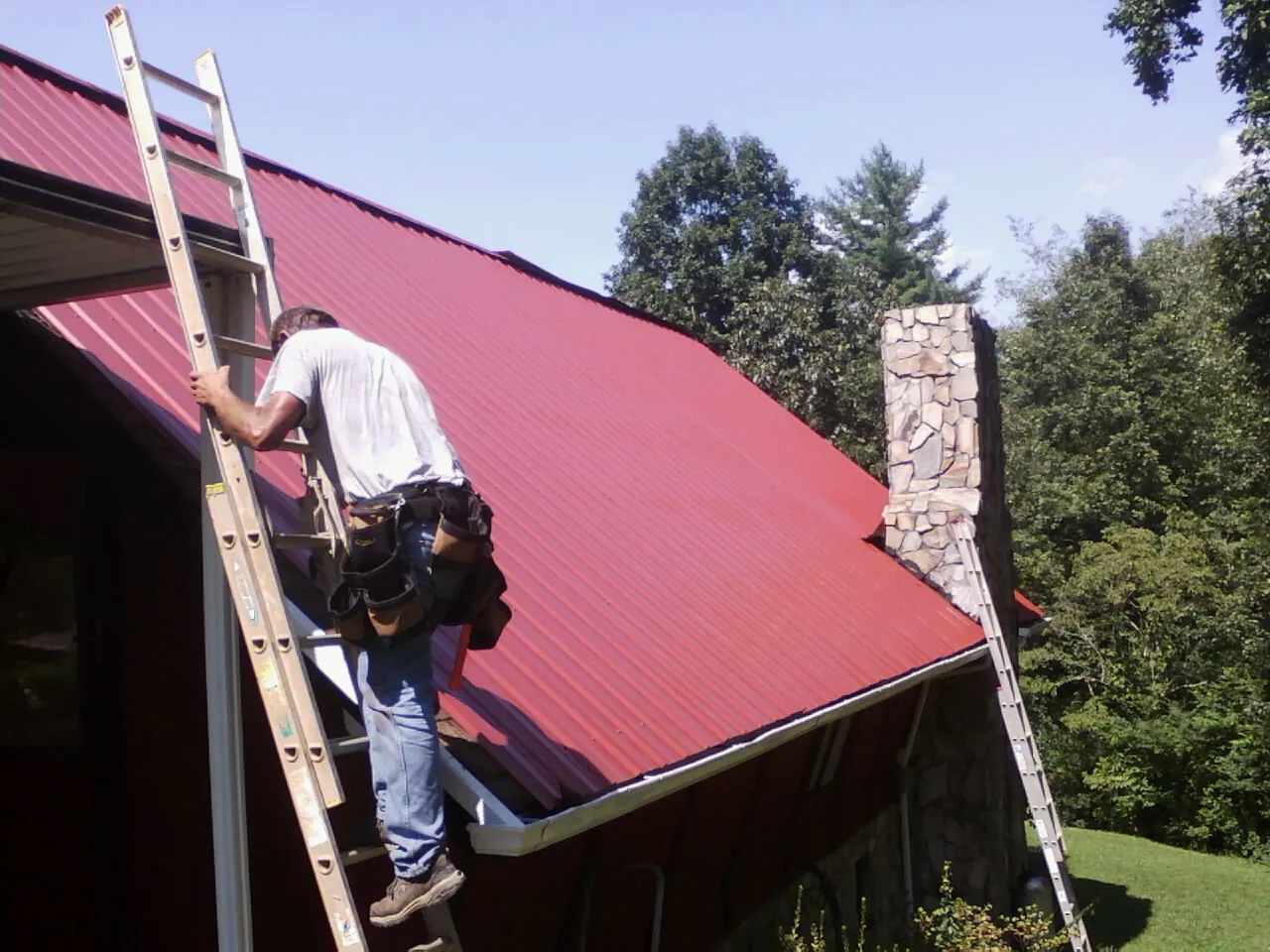 Roofer on a ladder beside a red metal roof and stone chimney — Soffit & Fascia Repair by Roofing Brothers in Wauneta